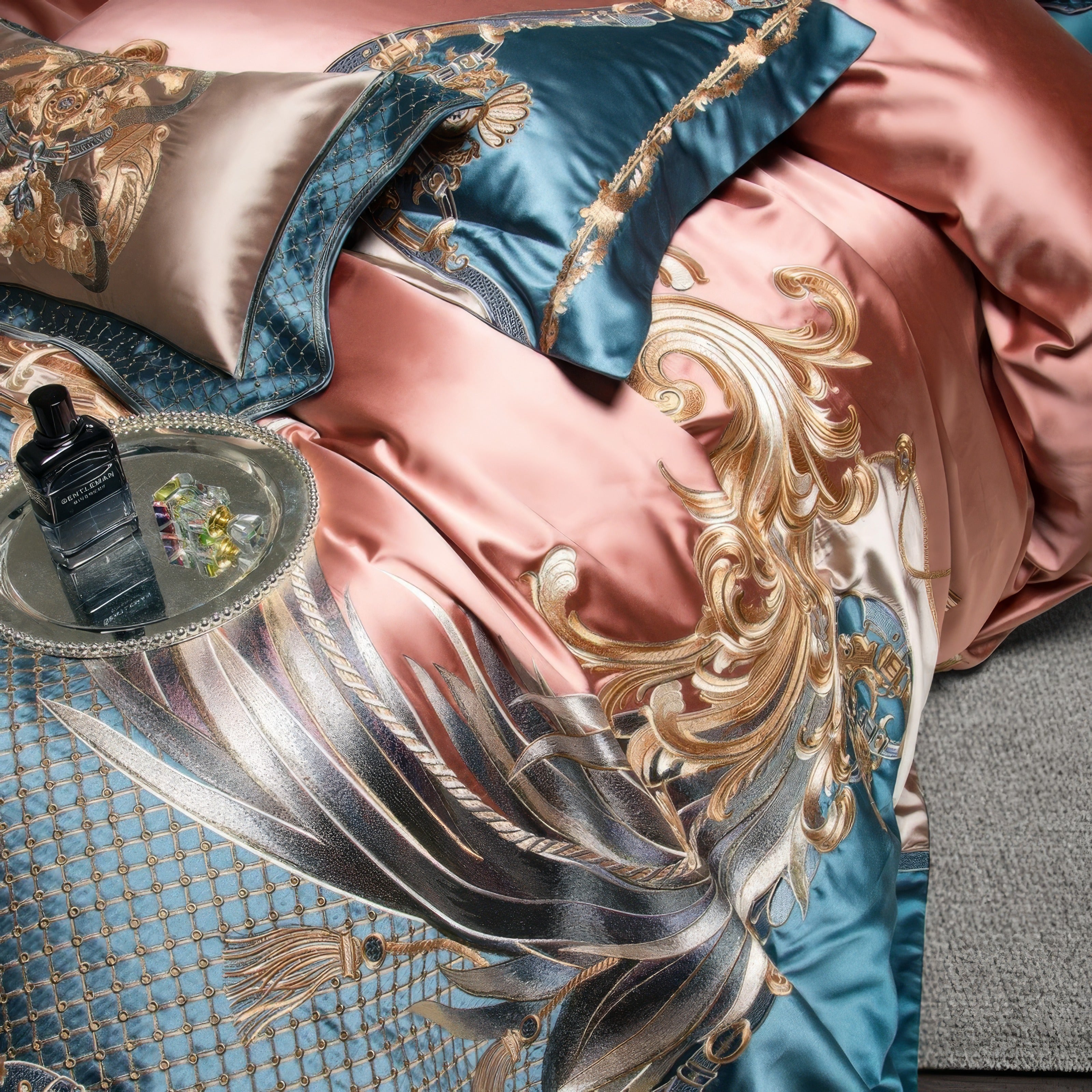 A close-up view of a luxurious bedding set adorned with intricate golden baroque patterns on a silky satin surface. The design features a harmonious blend of rose gold and teal hues, highlighted by metallic accents and ornamental swirls. A silver tray with a sleek black perfume bottle and vibrant decorative pieces rests atop the bedding, enhancing the lavish ambiance. The fabric's lustrous texture and elaborate detailing reflect opulence and refined craftsmanship.