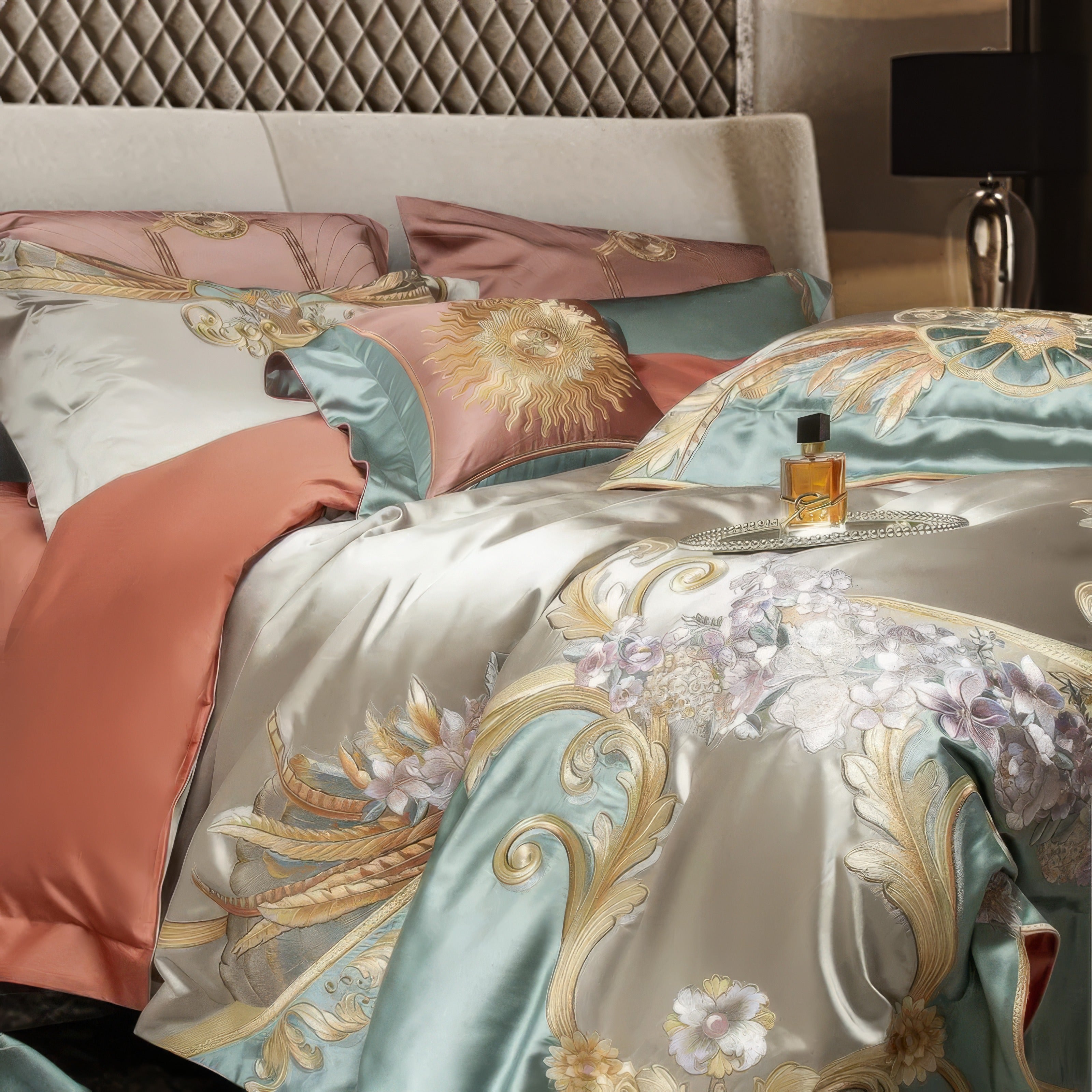 A close-up of a luxurious bedding set featuring intricate golden embroidery and soft floral patterns on a mint green and champagne satin fabric. The ensemble includes accent pillows in terracotta and sage, adorned with unique designs like a radiant sun motif. A bottle of perfume and a delicate chain on the bed add a personal, refined touch, set against the backdrop of a quilted headboard and modern decor.