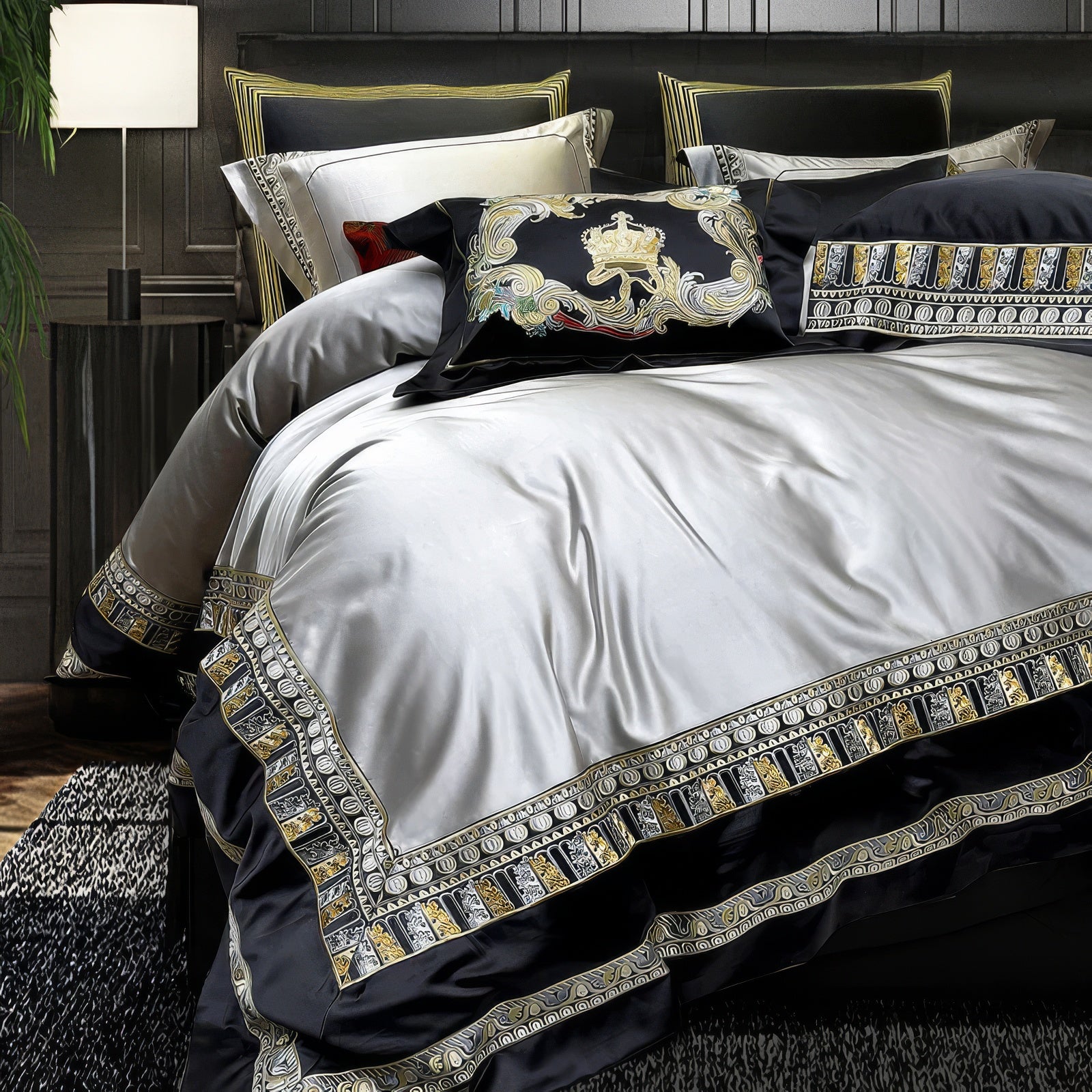 A close-up of an opulent silver and black bedding set featuring an intricately embroidered border in gold and black geometric and floral patterns. The centerpiece includes a decorative black velvet pillow with an ornate embroidered crest in gold, silver, and red, adding a regal touch. The smooth and lustrous fabric reflects a luxurious sheen, complementing the modern dark interior and textured carpet beneath.

