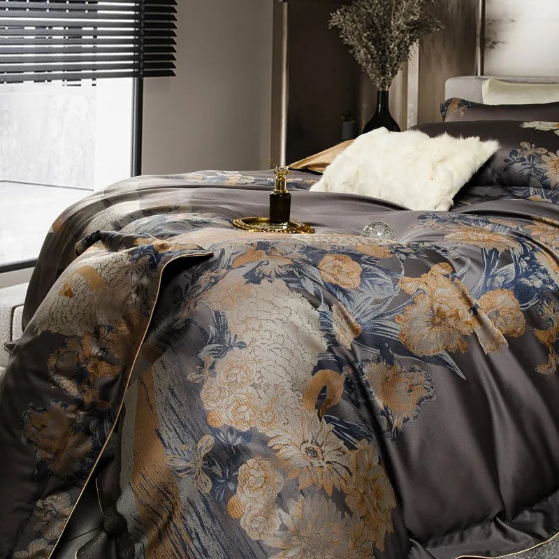 A side view of a luxurious dark gray bedding set adorned with gold and blue floral embroidery featuring swans. The detailed design flows seamlessly over the fabric, accentuated by a subtle sheen. A white faux fur pillow adds contrast, while a gold tray holding a perfume bottle and a decorative glass sphere enhances the opulent ambiance. A modern interior with natural light and a vase with dried branches completes the setting.