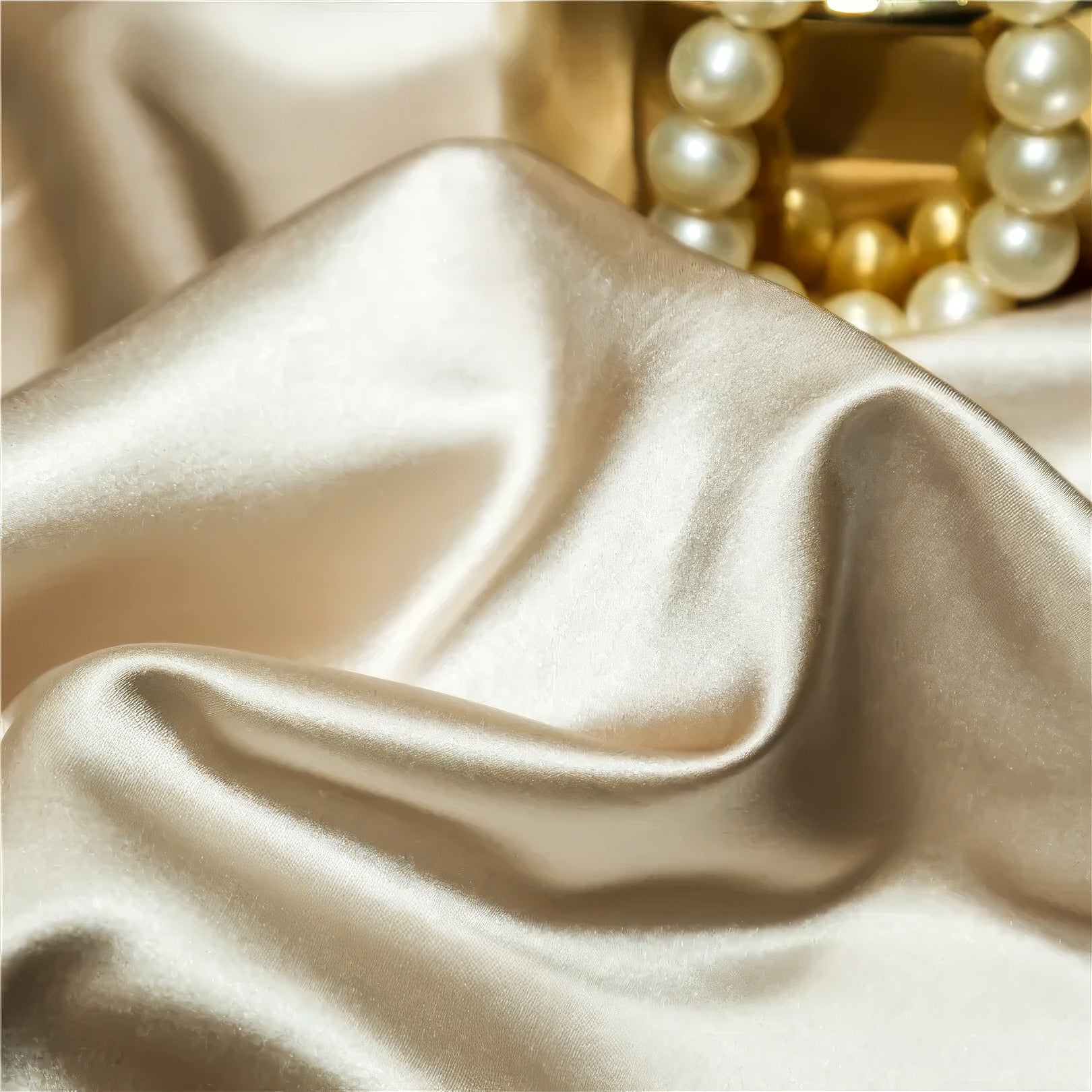 Close-up of luxurious beige silk fabric with a smooth, glossy texture, draped elegantly next to a decorative gold jar adorned with a string of pearls.

