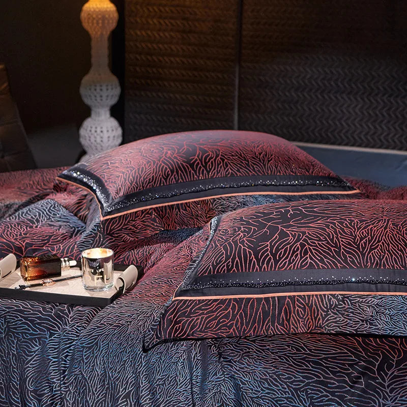 Close-up of two black and red pillows with coral-inspired patterns on a matching bedding set. A tray with a candle and small perfume bottle sits on the bed, with a textured wall and modern lamp in the background.

