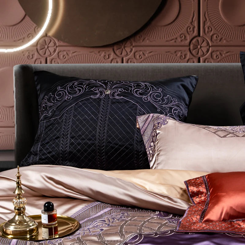 A detailed view of luxurious bedding featuring a black satin pillow with intricate embroidered patterns in silver and lavender, creating a sophisticated and regal look. The foreground showcases layers of soft, silky sheets in champagne, lavender, and coral hues. A vibrant coral-orange decorative pillow with diamond quilting and silver detailing adds a pop of color. The bedding is adorned with elegant embroidery and ornate patterns, complemented by a brass tray holding a small bottle of perfume and a decorat