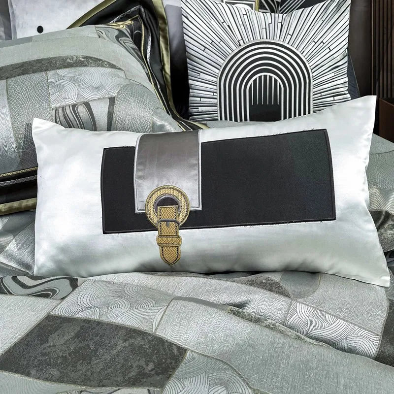 Decorative lumbar pillow from the Art Deco bedding set, featuring a bold black and silver design with a central gold embroidered buckle accent. Behind it is another pillow showcasing a black-and-white geometric pattern reminiscent of classic Art Deco architecture. The silky texture of the fabric enhances the luxurious appeal, complementing the sophisticated patchwork design of the bedding.