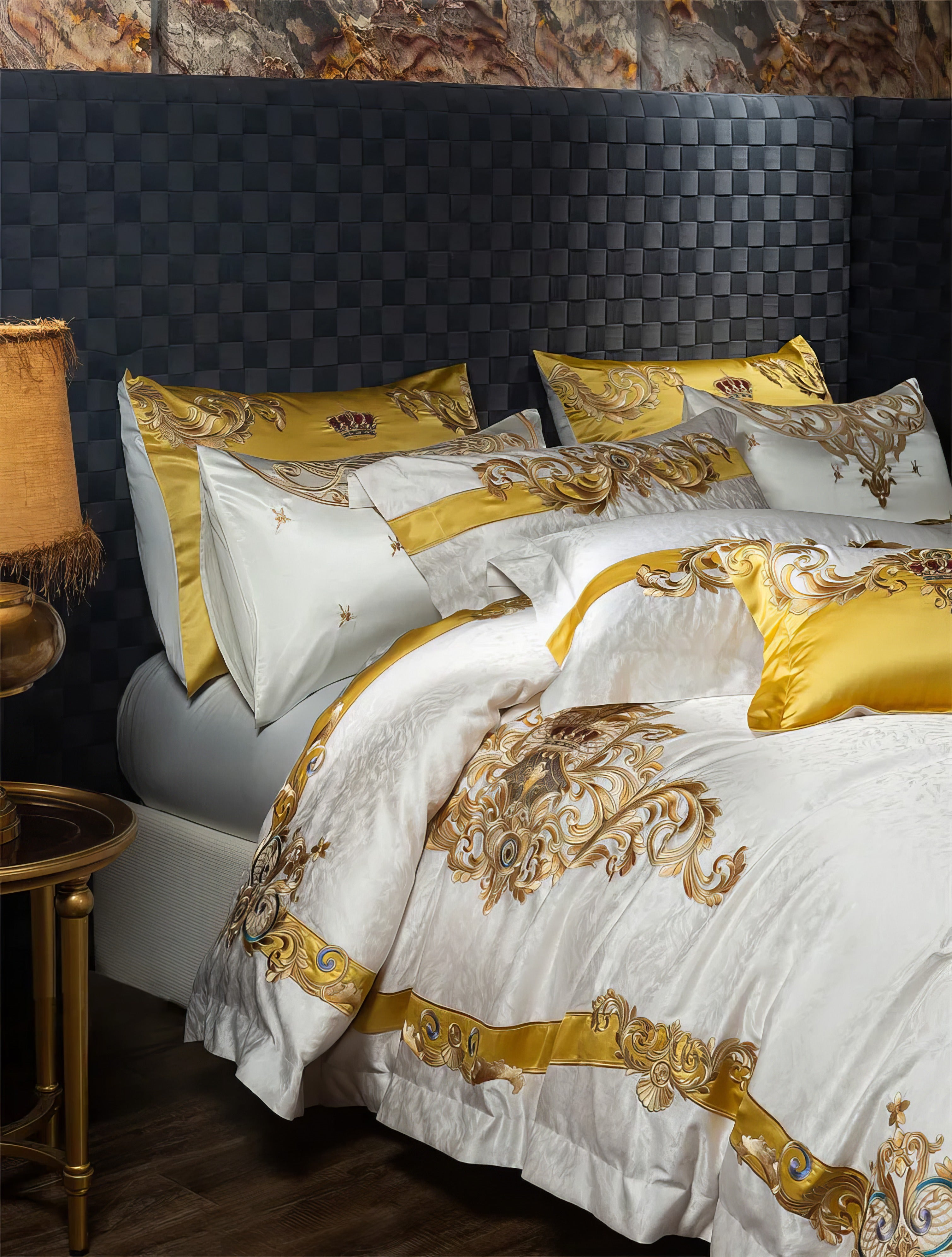 A luxurious bedding set with exquisite golden embroidery on a smooth white base, highlighted by ornate baroque designs and regal crown motifs. The set includes matching pillowcases and a vibrant golden accent pillow, framed against a sophisticated dark woven headboard. The warm tones of the gold detailing are complemented by a vintage-style lamp on a brass side table, creating an elegant and opulent bedroom atmosphere.