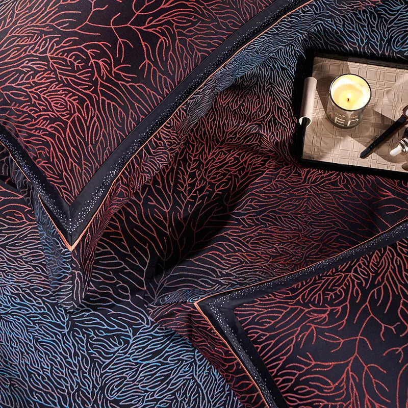 Close-up of black bedding with intricate red coral-inspired patterns, highlighting matching pillows and a small tray holding a lit candle, pen, and perfume bottle.

