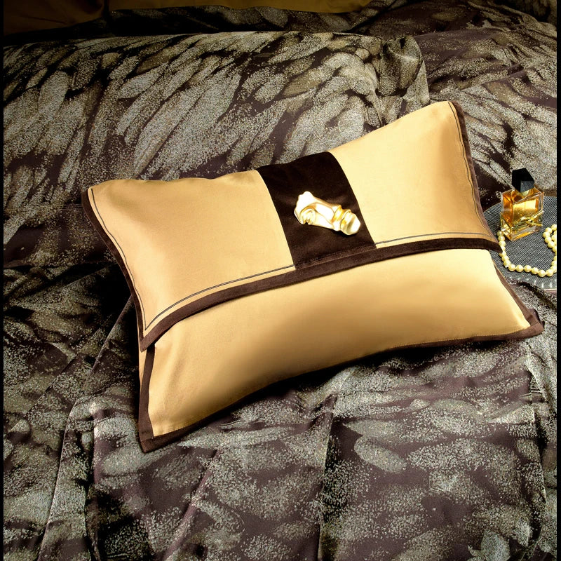 A rectangular decorative pillow in luxurious gold and brown hues, featuring a centered velvet accent strip with an intricate gold embellishment, resting atop a bedding set with a dark, abstract textured pattern. The pillow exudes sophistication and complements the surrounding opulent design elements, creating a visually striking addition to a lavish bedroom setup.