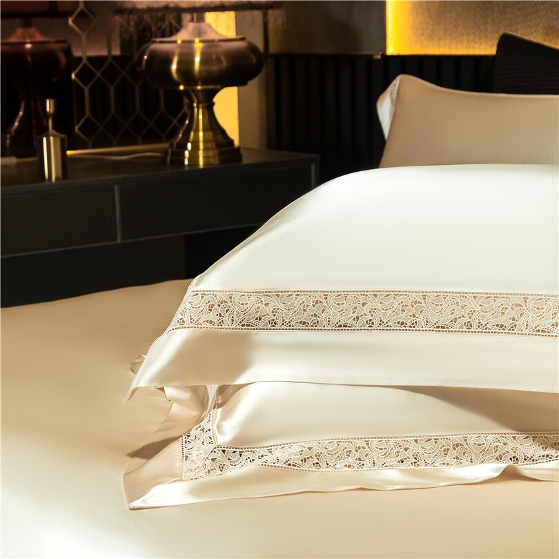 Close-up of luxurious cream-colored pillowcases adorned with delicate lace embroidery along the edges. The silky texture of the fabric reflects the ambient lighting, adding a soft glow. A golden table lamp and geometric wall accents create a warm, elegant backdrop, emphasizing the opulence of the bedding design.