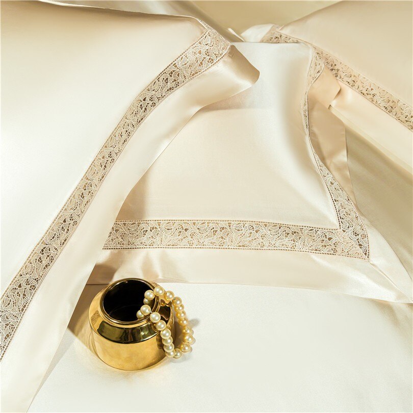 A close-up view of cream-colored silk pillowcases featuring intricate lace embroidery along the borders, showcasing their luxurious and elegant design. The glossy fabric reflects a soft sheen, enhancing its opulence. A small golden vase with a pearl bracelet sits nearby, adding a touch of sophistication to the composition.