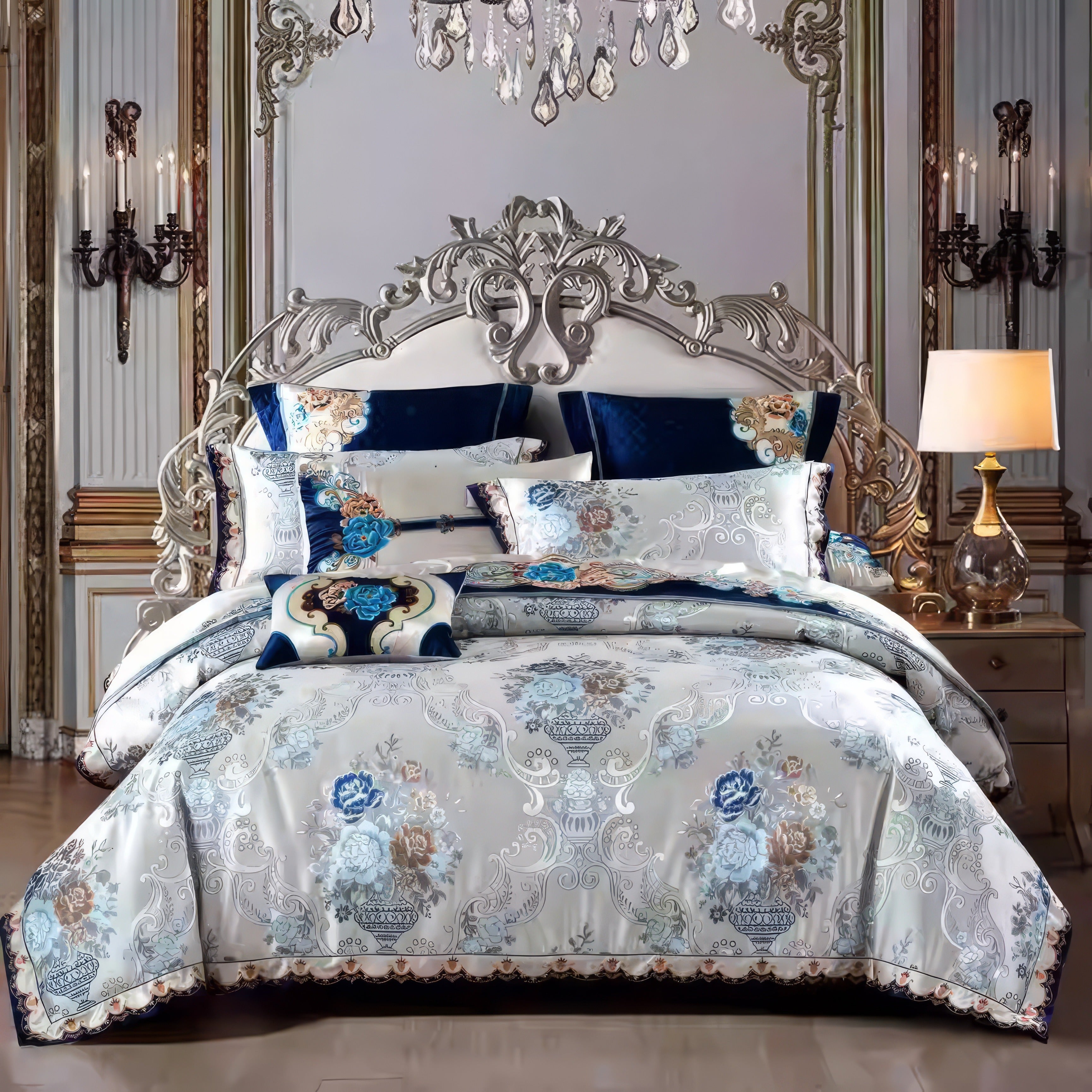 A regal bedroom adorned with an ornate metallic headboard featuring intricate curves and details, complemented by a luxurious bedding set. The bedding showcases a lavish floral design in shades of blue, gold, and cream, paired with accents of deep navy velvet pillows. The room exudes opulence, enhanced by crystal chandeliers and elegant wall-mounted candelabras. A gold-toned lamp on a classic side table adds a warm glow, completing this sophisticated and aristocratic ambiance.

