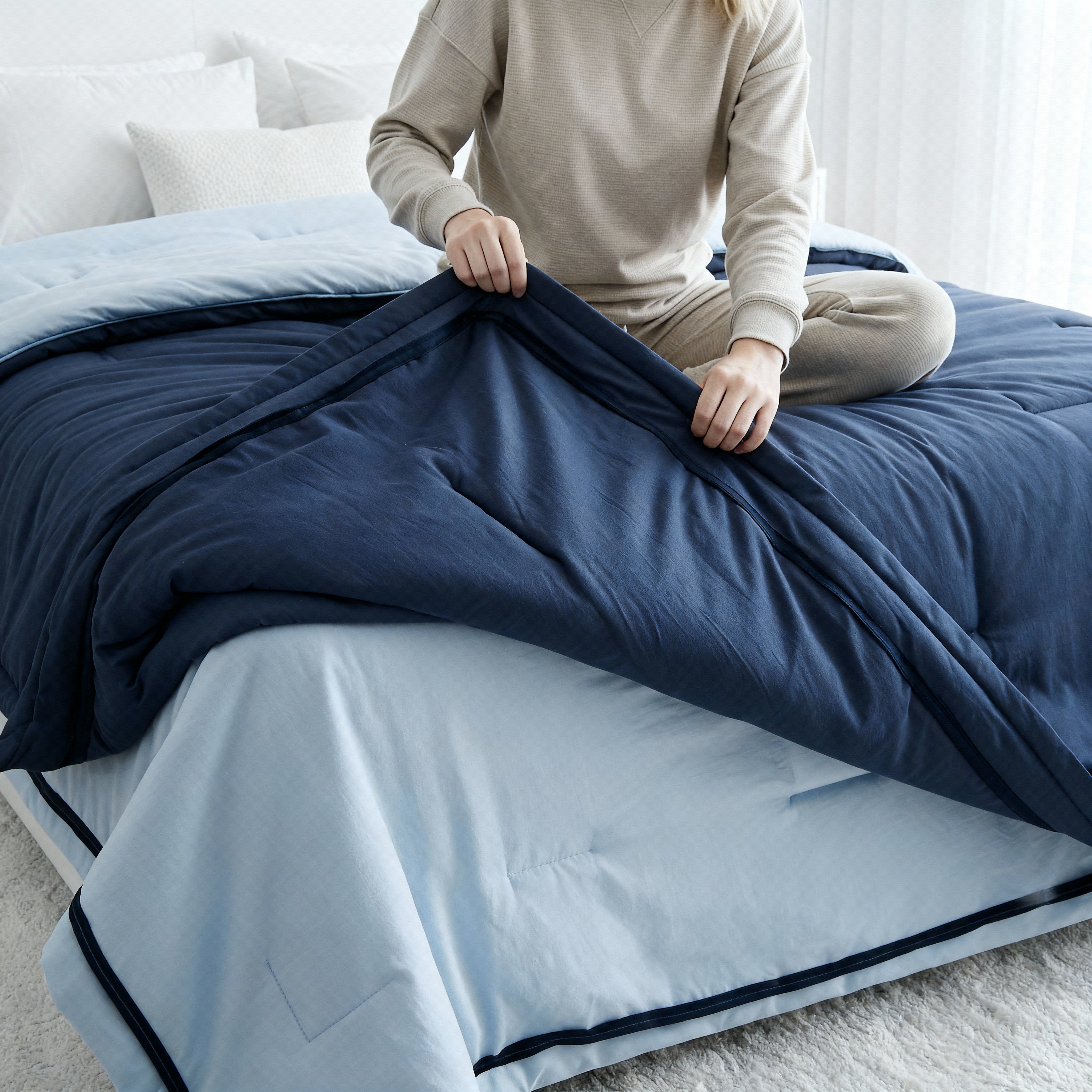 Person folding a blue comforter on a bed in a bright room.