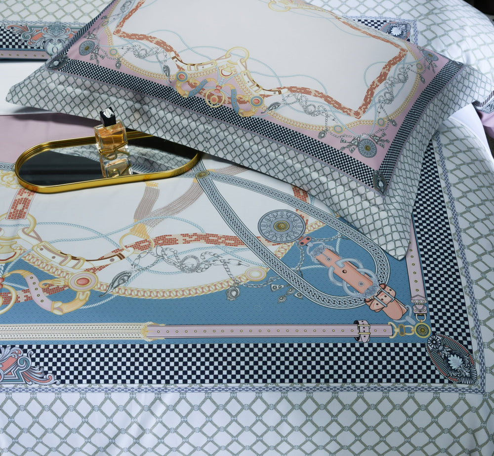 A detailed view of a bedding set with an equestrian-inspired design, featuring ornate reins, chains, and geometric patterns in pastel pink, blue, and white tones. A matching pillowcase with coordinating designs rests on the bed. A gold tray with a perfume bottle sits on the duvet cover.

