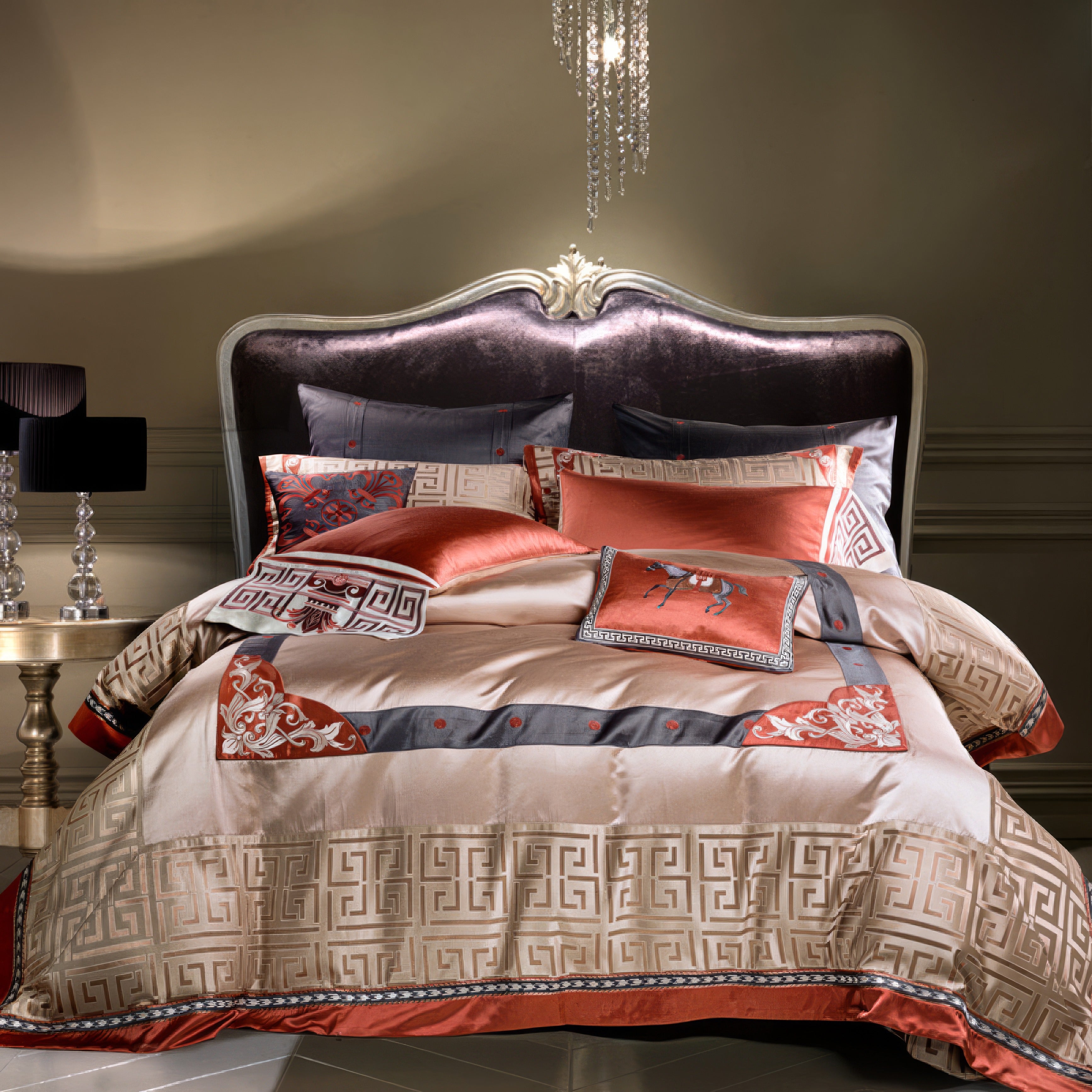 A luxurious bedding set featuring a mix of beige, gray, and vibrant orange tones with intricate Greek key patterns and embroidery accents. The plush headboard in a dark velvet material adds elegance, with a crystal chandelier above and a reflective side table adorned with a modern black lamp nearby.