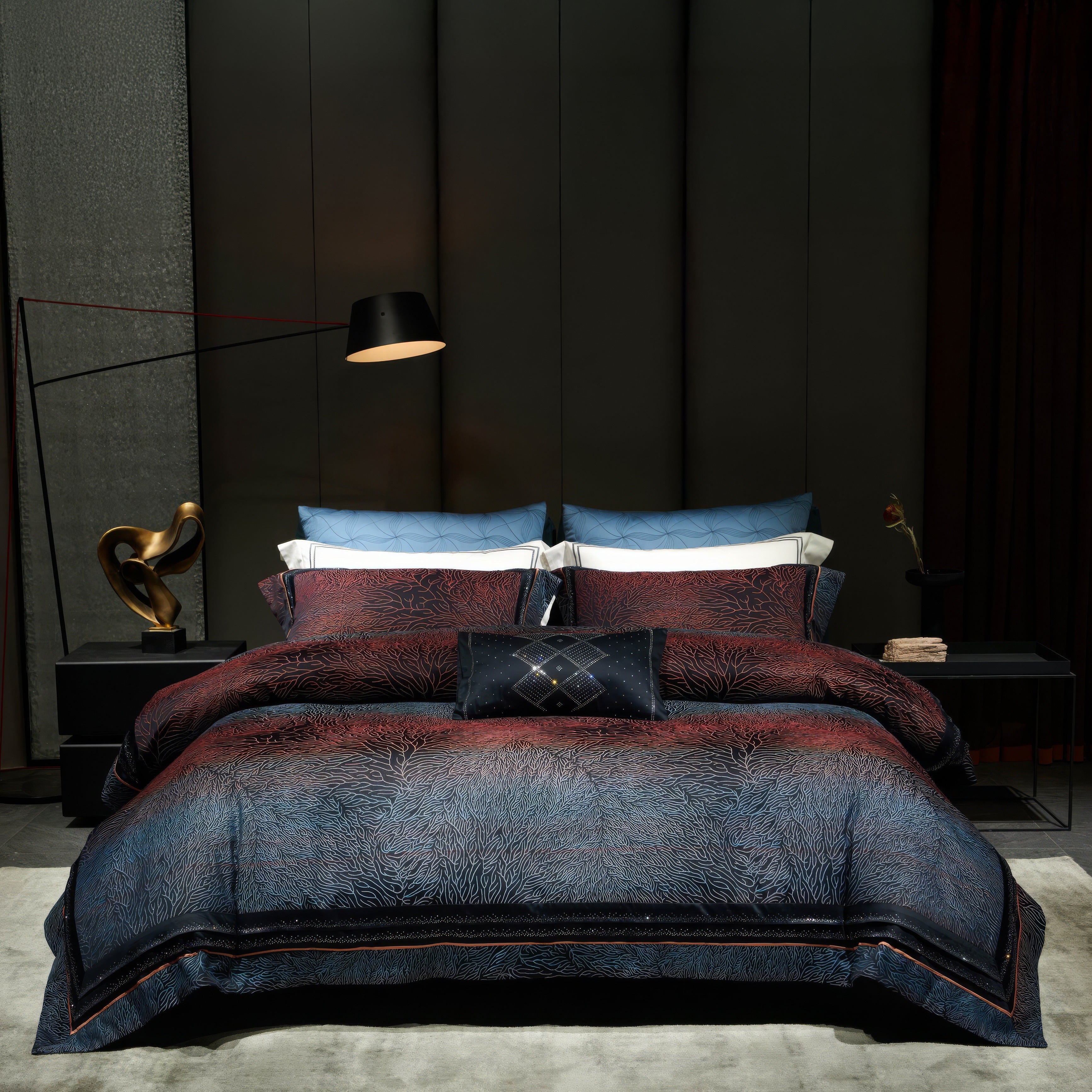 Luxurious bedding set in shades of deep red and navy blue with intricate, subtle patterns resembling coral textures, complemented by matching pillowcases and a decorative navy cushion with embroidered diamond detailing. The bedding sits against a modern, dark-paneled wall with a soft grey area rug beneath. A sleek black floor lamp and sculptural gold decor piece add sophistication to the room.

