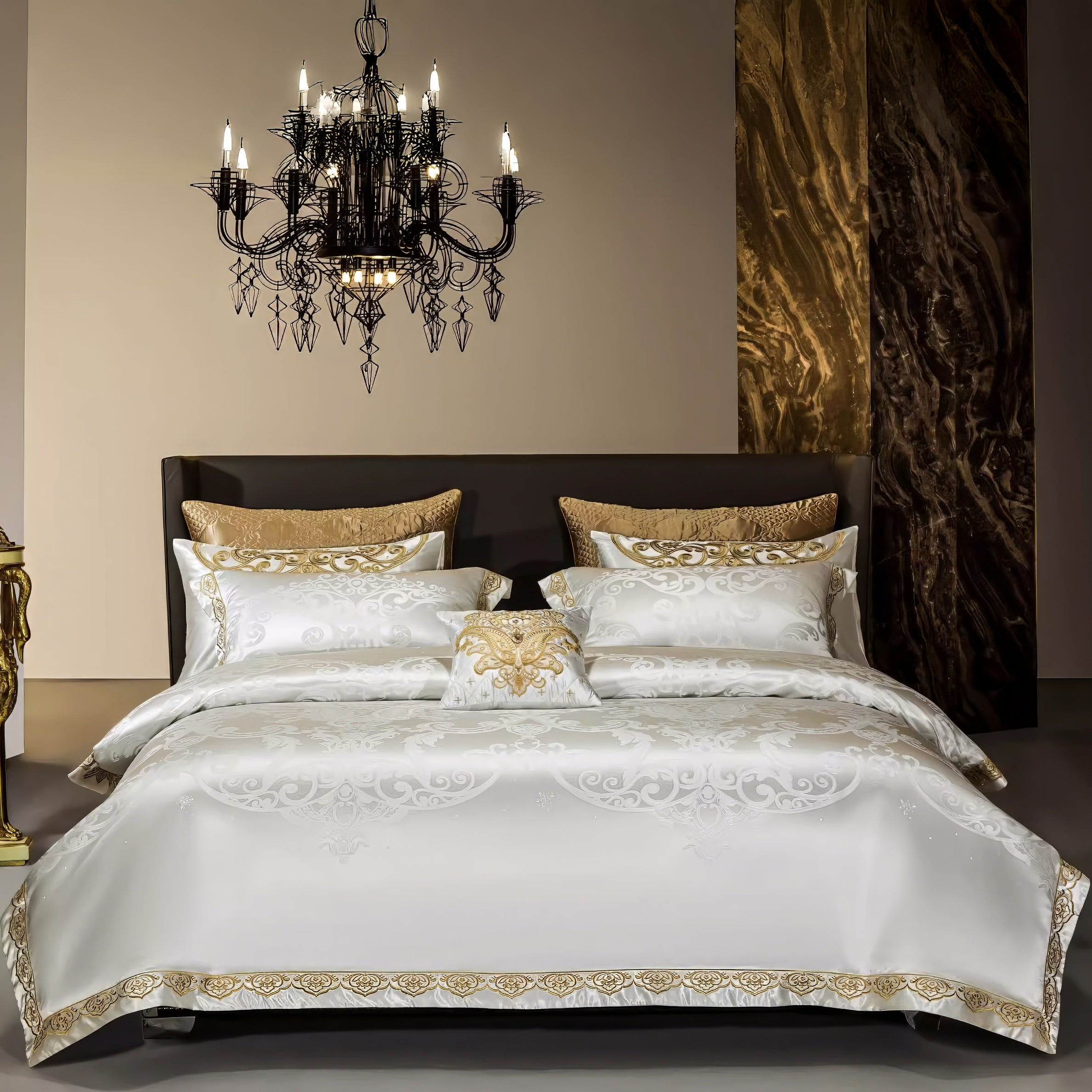 A luxurious white and gold bedding set with intricate embroidery, presented in a sophisticated setting. The bed is adorned with a matching decorative pillow, flanked by oversized golden accent pillows. The backdrop features a rich marble panel and a statement black chandelier with ornate details, enhancing the elegance of the room.

