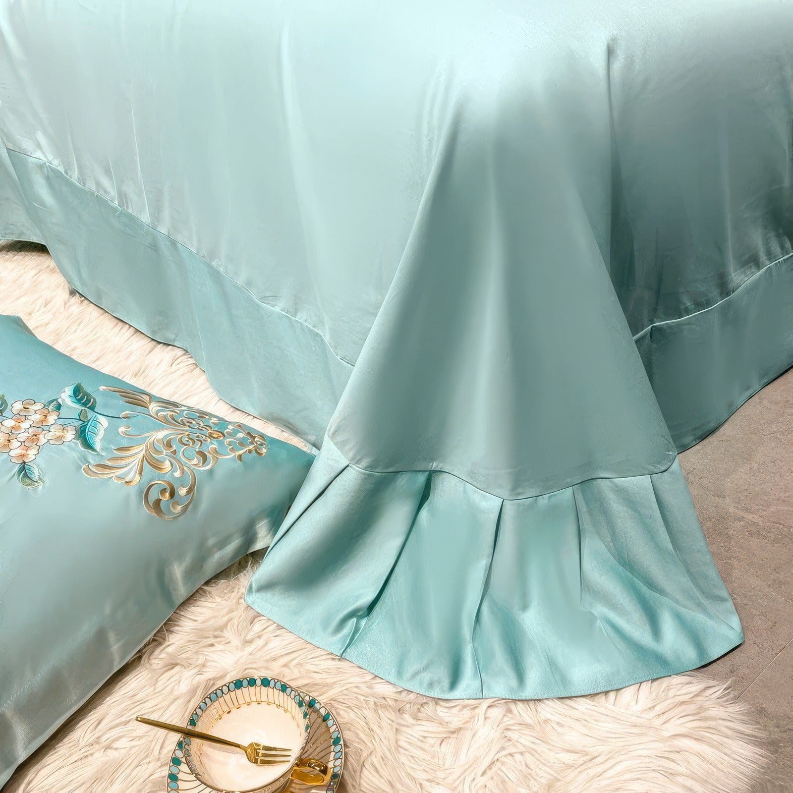 A close-up of an aqua blue bed skirt with an elegant draped design, paired with a decorative pillow showcasing intricate floral embroidery in gold and white tones. The setup is complemented by a ceramic plate with a gold fork resting on a fluffy white rug, adding a touch of sophistication to the scene.

