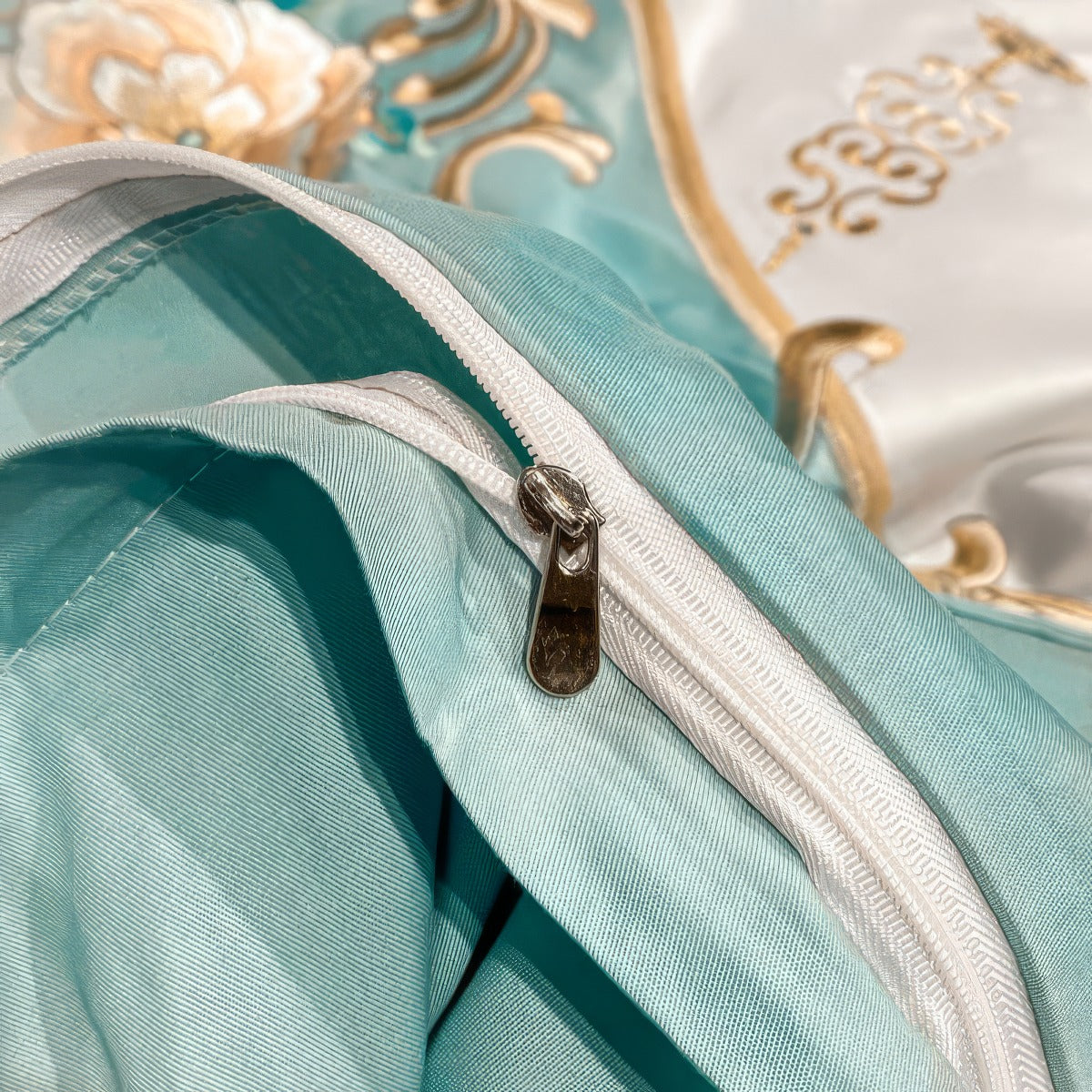 A detailed view of the aqua blue fabric featuring a durable white zipper closure, highlighting the fine stitching and smooth texture. The background includes elegant gold and white floral embroidery, adding a luxurious touch.

