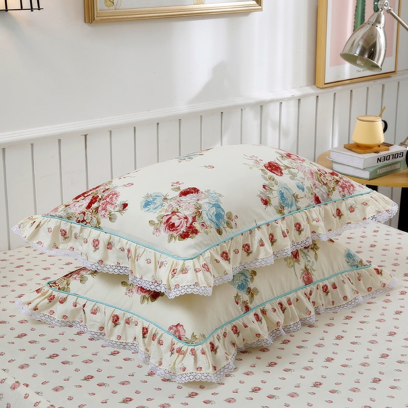 Two cream-colored pillowcases with floral designs featuring red, pink, and blue roses, accented by green leaves and small scattered blossoms. The edges are adorned with delicate ruffled trim and white lace, and the pillowcases rest on a matching bedspread with a small red floral pattern. A wooden nightstand in the background holds a lamp, a stack of books, and a cup.

