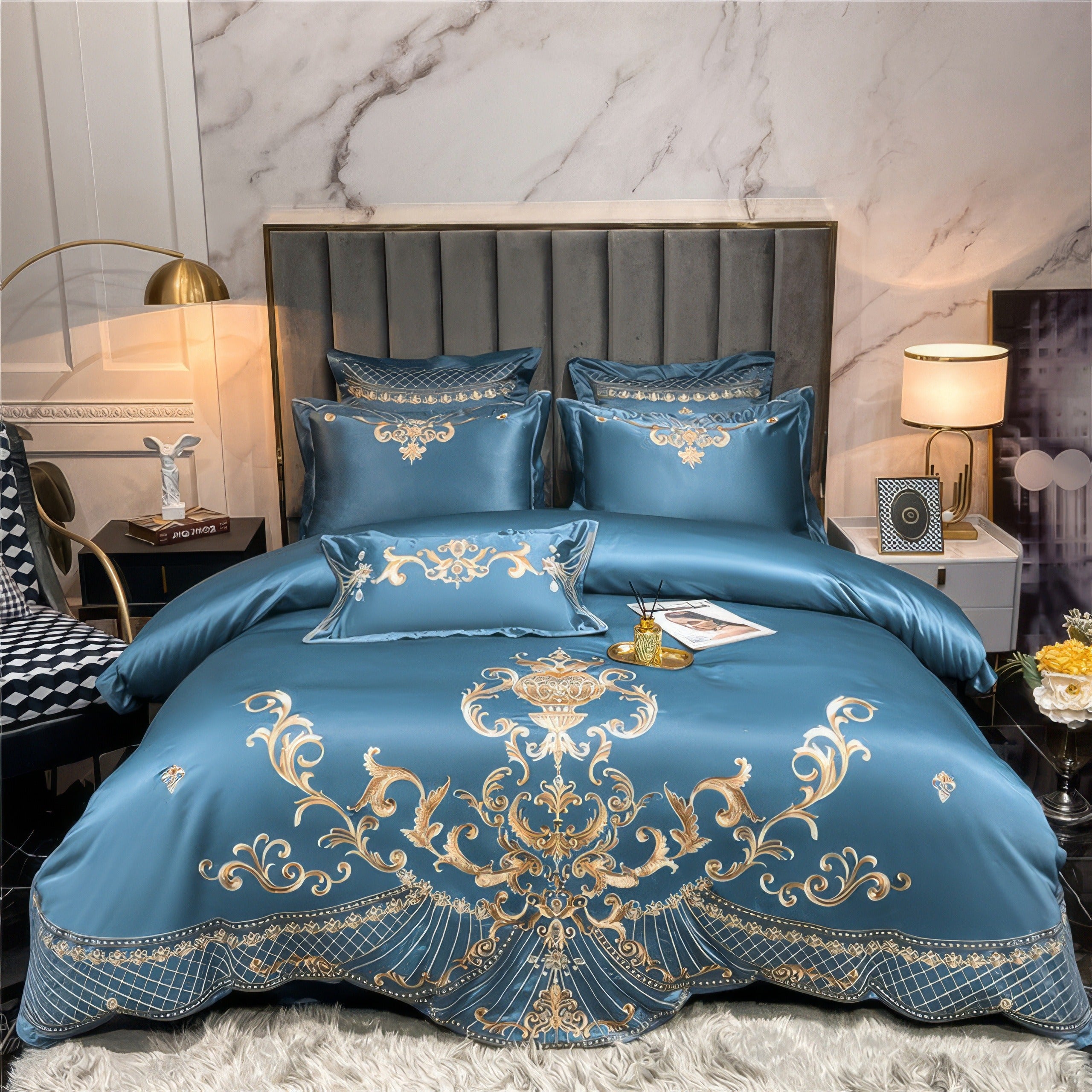 A luxurious blue bedding set featuring gold embroidery with intricate baroque-inspired patterns, matching pillowcases, and a decorative pillow, all styled on a contemporary bed with a grey upholstered headboard. A cozy white fur rug lies beneath the bed, and modern lamps with gold accents light up the space.

