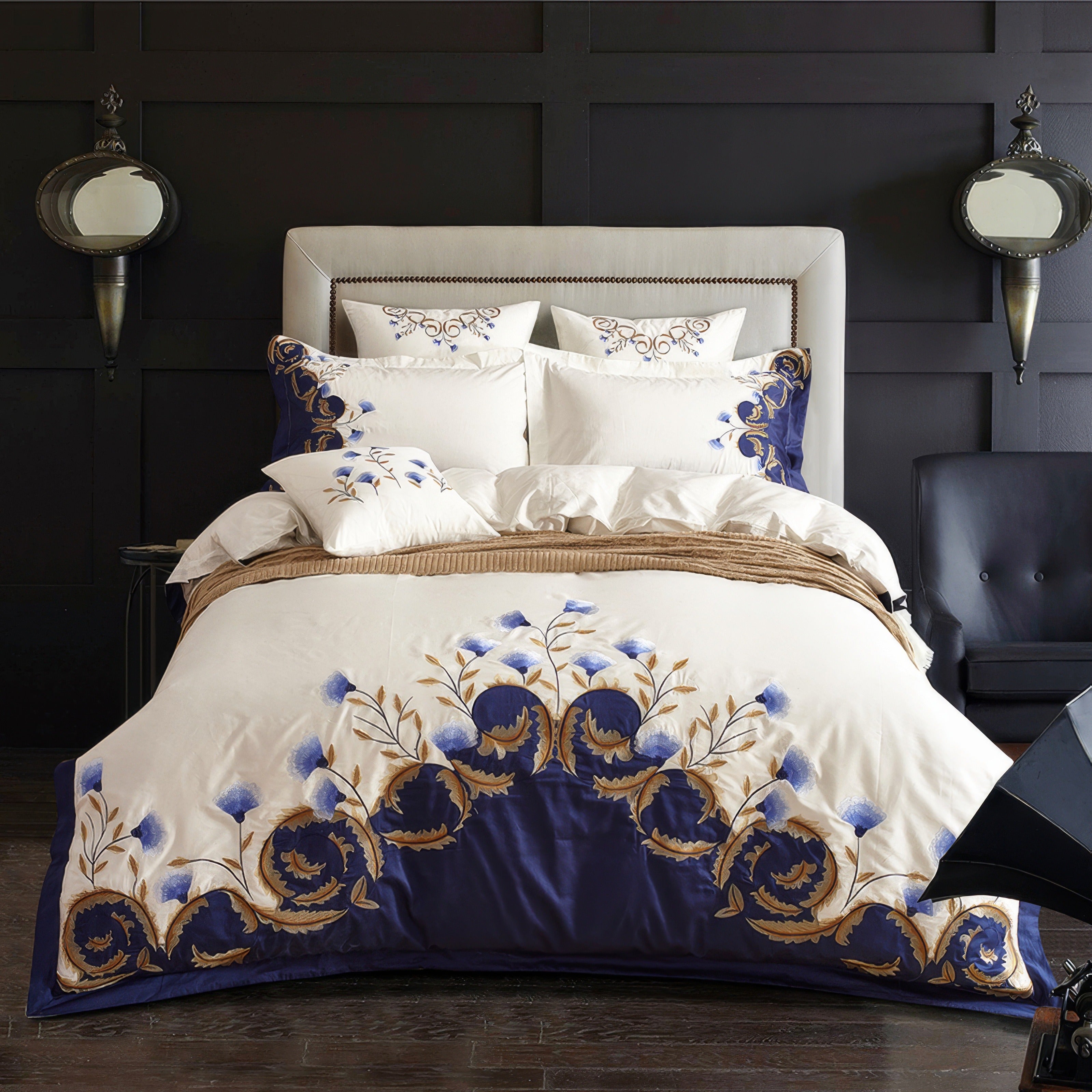 Egyptian cotton bedding set with intricate blue and gold floral embroidery on a white duvet cover, complemented by a navy-blue border. The set includes matching pillowcases with coordinating embroidery, arranged on a plush, upholstered headboard in a modern bedroom. The luxurious Egyptian cotton fabric enhances the softness and elegance of the bedding.

