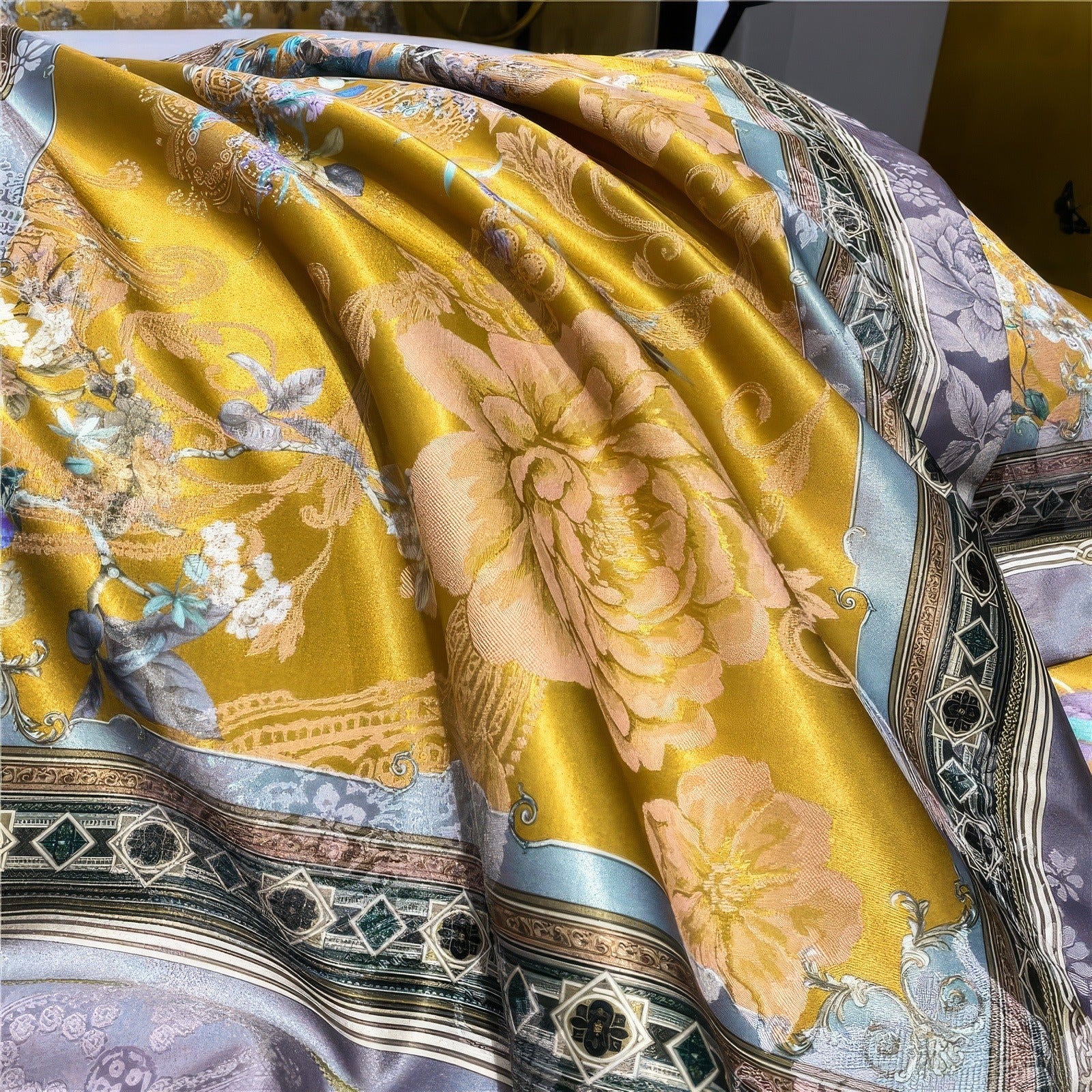 Close-up of a golden bedding fabric with intricate floral patterns, featuring large pastel flowers, ornate gray borders, and geometric accents in green and blue tones.

