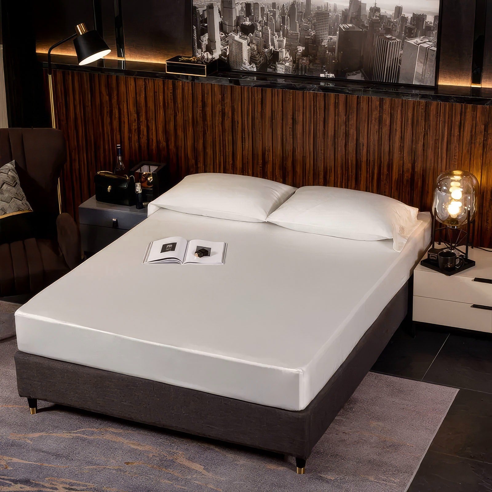 Minimalist bedroom with a sleek white fitted sheet on a modern bed frame, accompanied by two white pillows. The room features a dark wood-paneled accent wall, a contemporary nightstand with a glowing lamp, and decorative cityscape artwork in black and white. A small open book rests on the bed, enhancing the clean and modern aesthetic.

