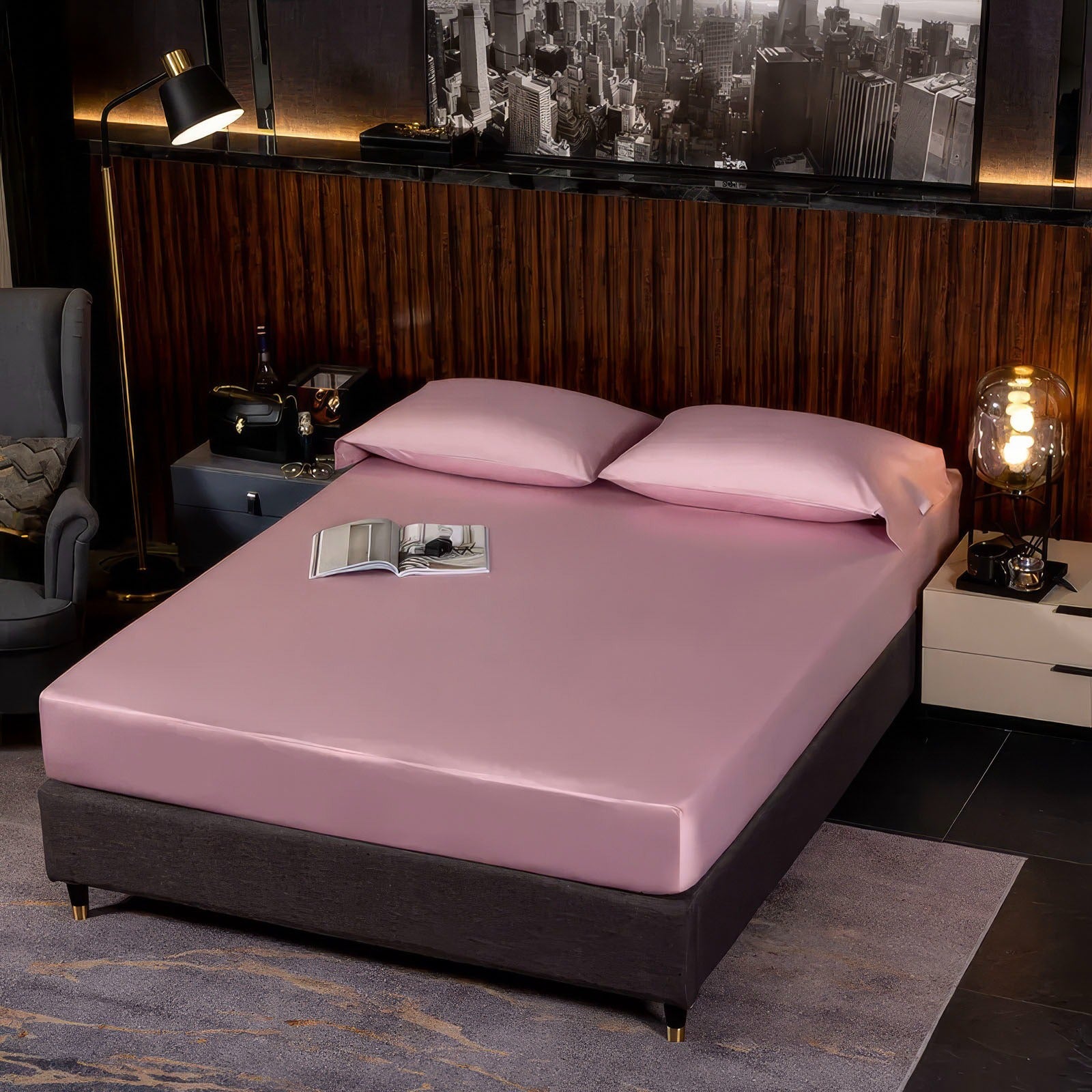 Modern bedroom with a soft pink fitted sheet and matching pink pillowcases on a sleek bed frame. A dark wood-paneled accent wall is paired with a black-and-white cityscape artwork. A lit, modern glass lamp on the nightstand adds a warm glow, while an open book rests on the bed, contributing to the room's cozy yet stylish aesthetic.

