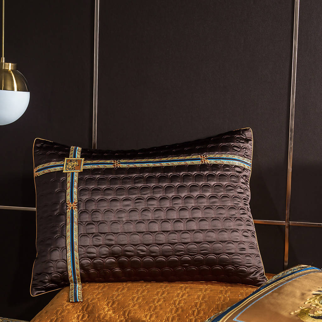 Dark brown decorative pillow with embossed circular patterns and ornate gold and blue ribbon detailing, featuring a small gold emblem accent, resting against a black background.

