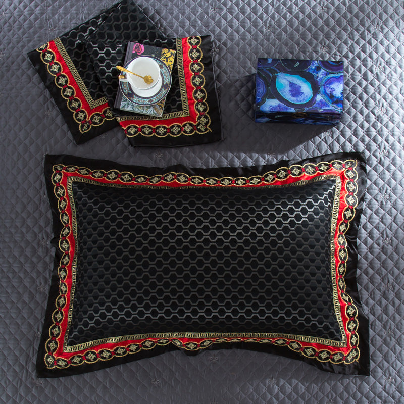 A black rectangular pillow with a geometric hexagonal pattern, bordered by an intricate red and gold baroque design. The pillow rests on a quilted gray bedspread adorned with subtle fleur-de-lis motifs. Nearby, matching decorative black and gold fabric is displayed, accompanied by a blue patterned box and an elegant teacup set on a tray, showcasing an opulent and cohesive bedding design.

