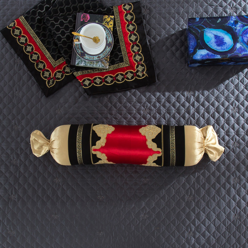 A cylindrical bolster pillow in black and gold with an ornate red center panel, featuring intricate gold embroidery and satin ribbon ends. The pillow is displayed on a quilted gray bedspread with subtle fleur-de-lis patterns, alongside matching fabric with gold and red baroque detailing, a decorative blue box, and a teacup set resting on an elegant tray.

