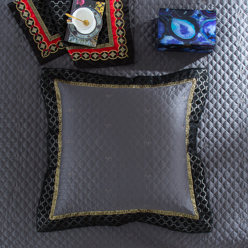 A square decorative pillow in luxurious gray fabric with a quilted pattern and subtle fleur-de-lis embroidery, framed by a black border with intricate gold and red detailing. The pillow rests on a matching quilted bedspread, while a vibrant blue box and an ornate black and red fabric with gold accents featuring a teacup complete the regal arrangement.

