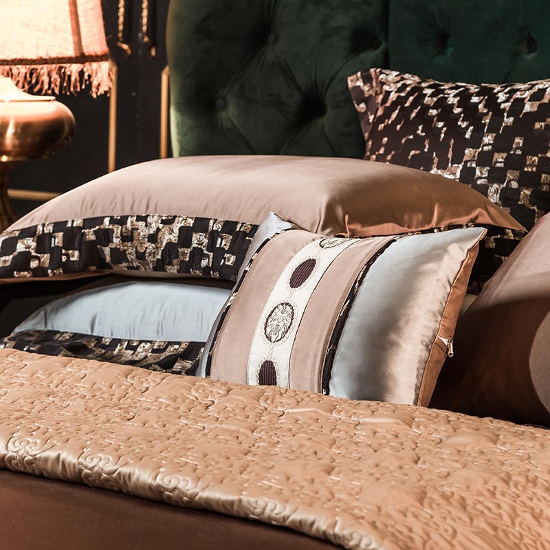 A close-up view of a luxurious bedding set with an elegant color palette featuring golden brown, light blue, and black. The decorative pillow in the center showcases intricate circular embroidery on a silky light blue and gold fabric, bordered with black patterned trim. Behind it, smooth satin pillowcases in golden brown and patterned black provide a rich, opulent texture. The quilted blanket in the foreground adds depth with its embossed floral design, completing the sophisticated, high-end aesthetic.

